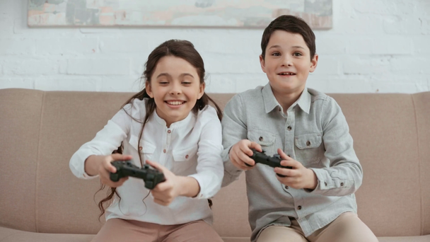 KYIV, UKRAINE -  APRIL 15, 2019: front view of two laughing kids sitting on sofa, holding joysticks and pushing each other while playing video game - Video