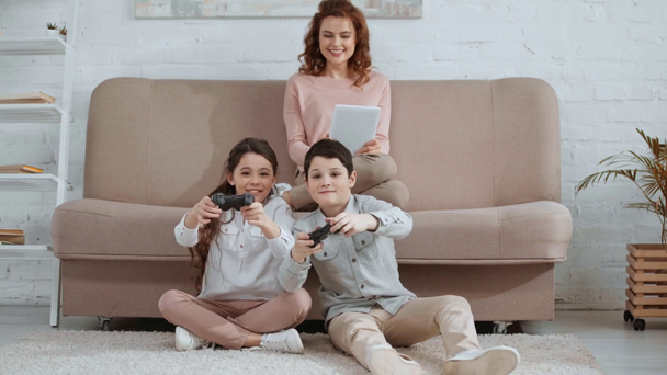 KYIV, UKRAINE -  APRIL 15, 2019: front view of smiling children with joysticks playing video game while mother sitting on sofa and using digital tablet - Πλάνα, βίντεο