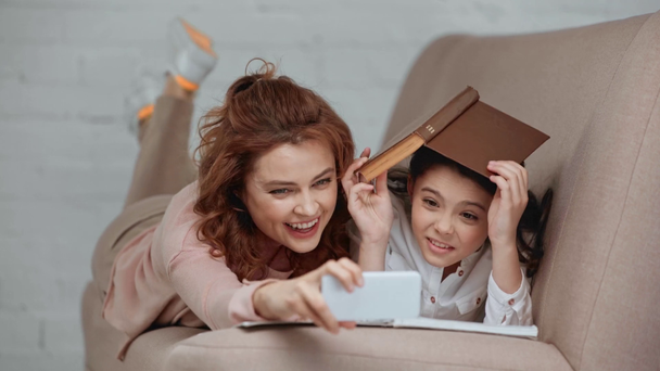 χαρούμενο Προεφηβικό παιδί βάζοντας το βιβλίο στο κεφάλι, παίρνοντας selfie με τη μητέρα και χαμογελώντας ενώ ξαπλωμένοι στον καναπέ στο σπίτι - Πλάνα, βίντεο