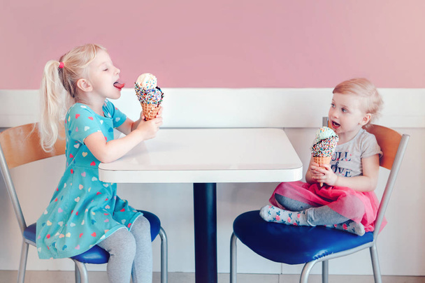 Lifestyle πορτρέτο των δύο χαρούμενος Καυκάσιος χαριτωμένο αξιολάτρευτο αστεία παιδιά κορίτσια κάθονται μαζί τρώγοντας γλείψιμο παγωτού με πολύχρωμα τρούφα. Η αγάπη ζηλεύει τη φιλία. Νόστιμο καλοκαιρινό φαγητό - Φωτογραφία, εικόνα