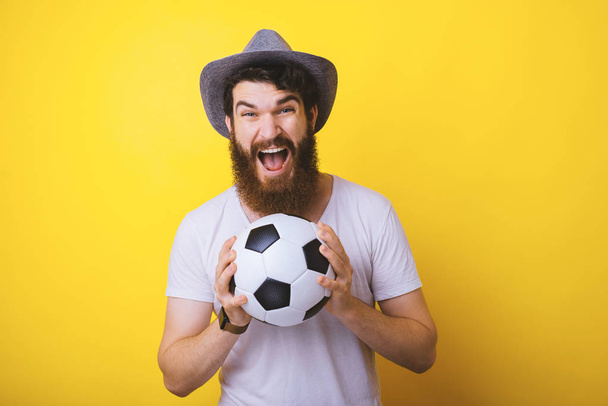 Excited Young bearded man catching a football or soccer ball on yellow background. - Photo, Image