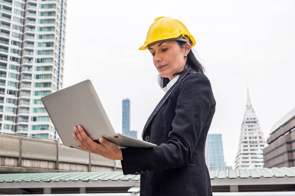 Woman engineer with yellow helmet holding laptop on urban backgr - Φωτογραφία, εικόνα