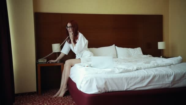 Kobieta siedzi na łóżku i rozmawia na telefon hotelowy - Materiał filmowy, wideo