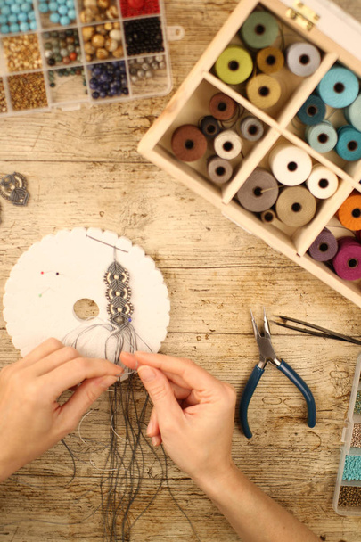 Κάτοψη των γυναικείων χεριών που φτιάχνουν ένα βραχιόλι με κουμιχίμο σε ξύλινο τραπέζι με εργαλεία, καρούλια από νήμα, φυσικές πέτρες και χρωματιστές χάντρες - Φωτογραφία, εικόνα