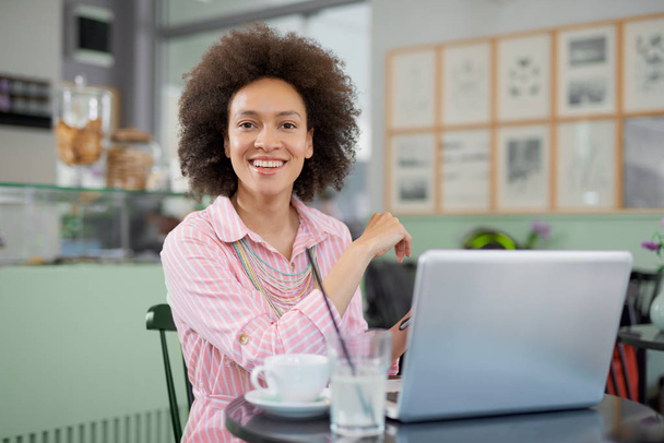 Gyönyörű mosolygó vegyes verseny nő ül a kávézóban és használata laptop. - Fotó, kép