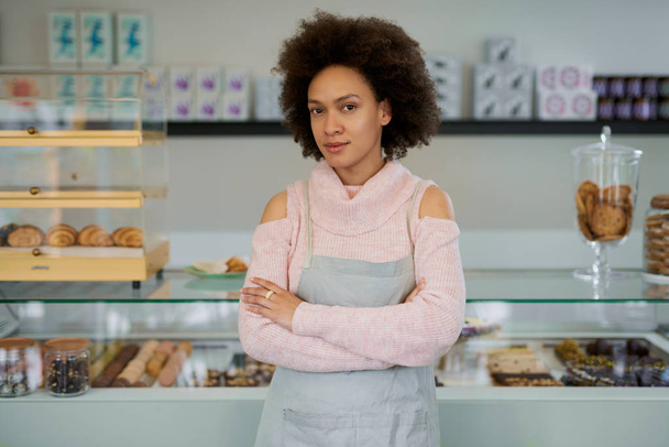 Mooie glimlachend gemengd ras vrouwelijke eigenaar van banketbakkerij gekleed in schort staande met armen gekruist in de winkel. - Foto, afbeelding