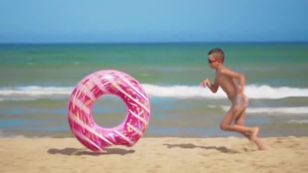 De jongen loopt langs het strand met een roze opblaasbare donut, rolt het langs het zand tegen de achtergrond van de zee. Het concept van ontspanning en plezier. - Video