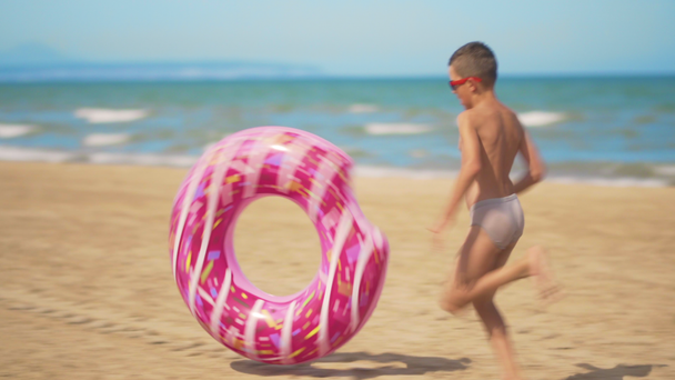 De jongen loopt langs het strand met een roze opblaasbare donut, rolt het langs het zand tegen de achtergrond van de zee. Het concept van ontspanning en plezier. - Video