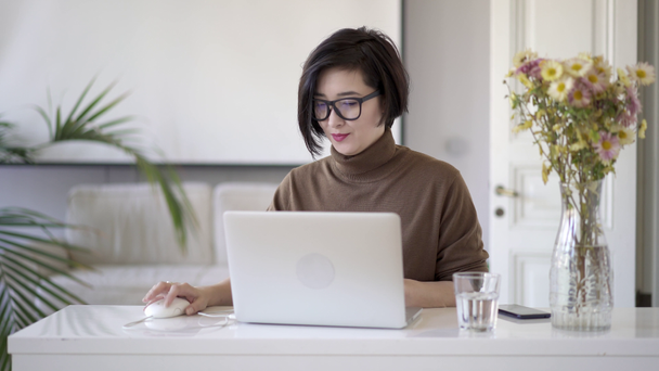 Asian woman in eyeglasses writing in laptop in white home office - Metraje, vídeo