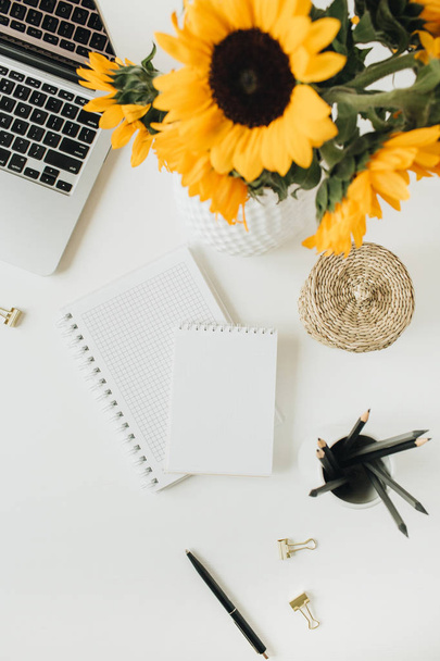 Flachbild-Schreibtisch mit Laptop, Notizbuch, gelbem Sonnenblumenstrauß auf weißem Hintergrund. von oben Freelancer / Blogger Sommer florales Arbeitskonzept. - Foto, Bild