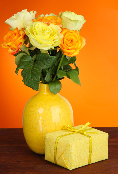 Beautiful bouquet of roses in vase with gift on table on orange background - Foto, Imagem