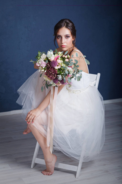 A young bride in a white dress sitting barefoot on a chair, with a bouquet of flowers posing against a blue wall - Fotó, kép