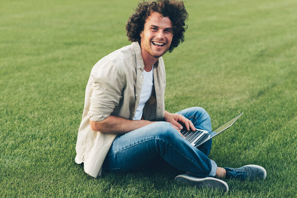 Image of happy young man with curly hair using laptop for chatting online with friends, connected to free wireless, sitting on the green grass. Handsome male typing on his laptop computer in the park - Fotó, kép