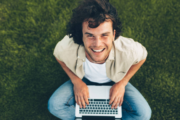 Overhead view of happy student sitting on the ground, working on laptop. Smiling man with curly hair using laptop for chatting online with friend, on the grass. Male typing on his computer in the park - 写真・画像