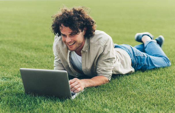 Cheerful young freelance man sitting on the ground, working on laptop. Smiling man with curly hair using laptop for chatting online with friends, on the grass. Male typing on his computer in the park - Фото, изображение