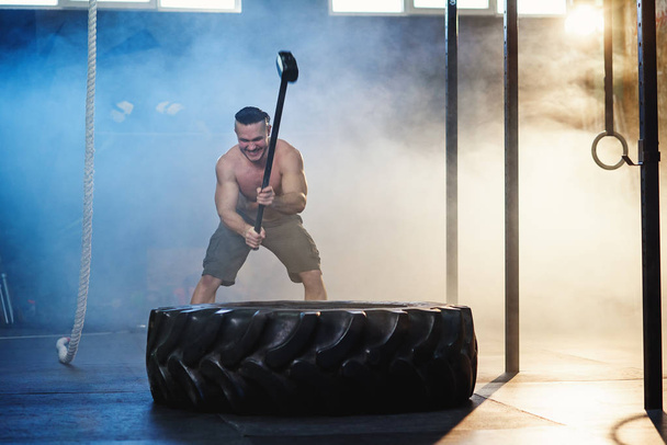 Attractive sport man hitting wheel tire with hammer during training. - Foto, afbeelding