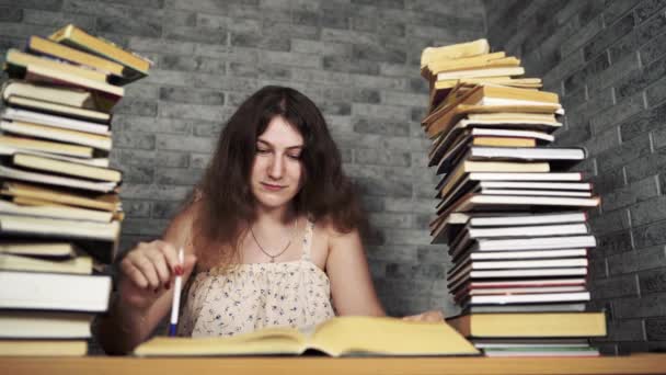 Tired female student reading among books. Pensive young woman sitting at table with pile of book and reading on background of grey wall - Materiaali, video