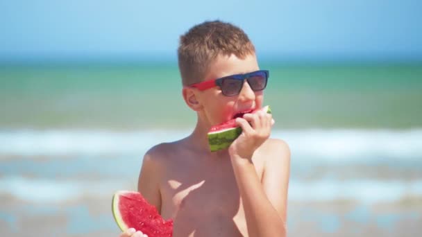 Το αγόρι με τα μαύρα γυαλιά τρώει καρπούζι στη θάλασσα. - Πλάνα, βίντεο