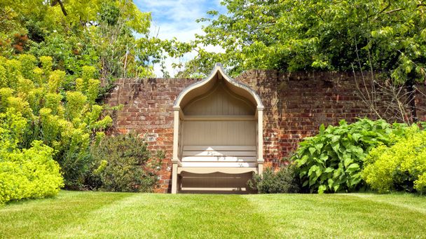 abgeschiedene Vintage-Holzbank an einer Ziegelmauer, in einem eleganten Landschaftsgarten, an einem sonnigen Tag in einer englischen Landschaft . - Foto, Bild