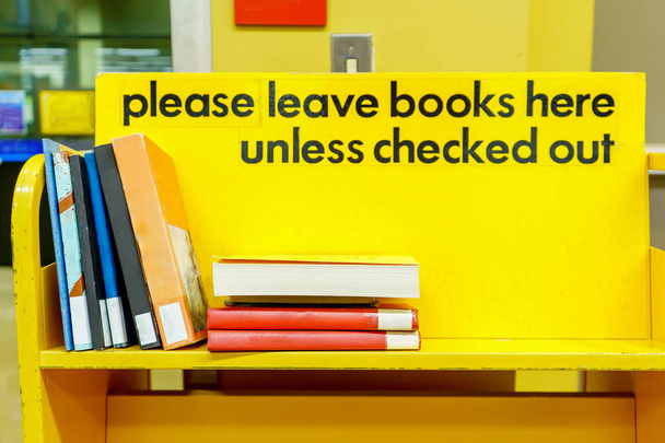 Gelber Bücherwagen mit Schild in schwarzen Buchstaben, und Bücher gestapelt in einem Regal zum Abstellen. - Foto, Bild