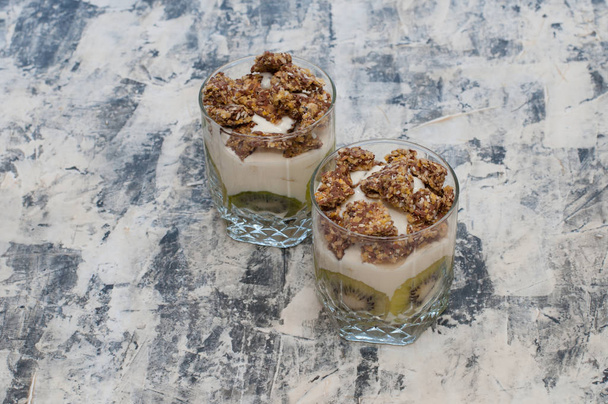 Гранола, ківі, банан та грецький йогурт у скляних чашках на сірому бетоні
 - Фото, зображення
