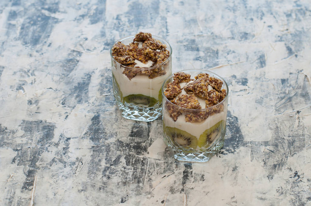 Granola, kiwi, banana and greek yogurt in glass cups on gray concrete - 写真・画像