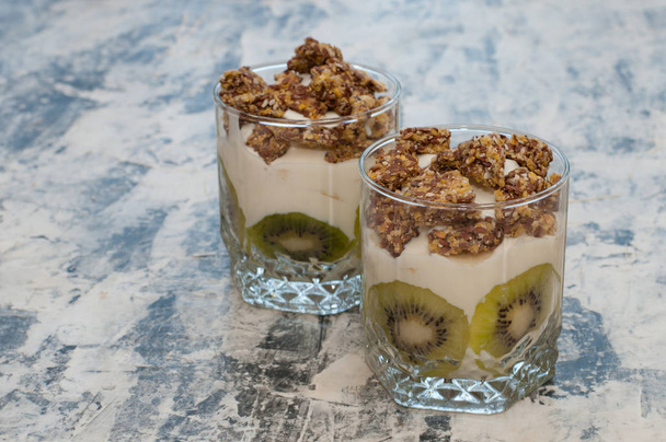 Granola, kiwi, plátano y yogur griego en dos tazas de vidrio sobre hormigón gris
 - Foto, imagen
