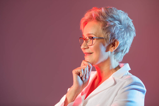 beautiful stylish woman aged, wearing glasses, in a white jacket on a pink background - Foto, imagen