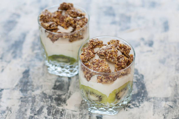 Granola, kiwi, banana and Greek yogurt in two glass cups on gray concrete, copy space. Fitness, figure, body and healthy food - Foto, Imagem