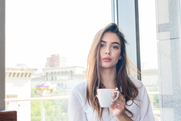 Een jong mooi bruin harige meisje in een wit shirt drinken cappuccino koffie - Foto, afbeelding
