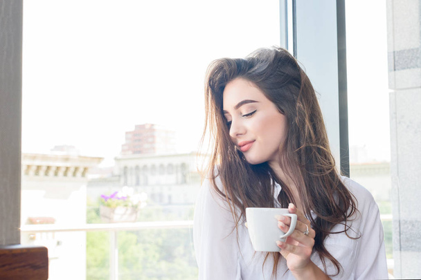 Nuori kaunis ruskeahiuksinen tyttö valkoisessa paidassa juomassa cappuccino-kahvia
 - Valokuva, kuva