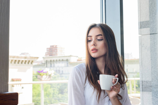 Молодая красивая темноволосая девушка в белой рубашке пьет кофе капучино
 - Фото, изображение
