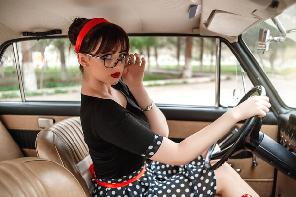 portrait of Caucasian beautiful young girl in black vintage dress and vintage glasses posing behind the wheel of a vintage car - Photo, Image