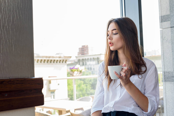ein junges schönes braunhaariges Mädchen in weißem Hemd trinkt Cappuccino-Kaffee - Foto, Bild