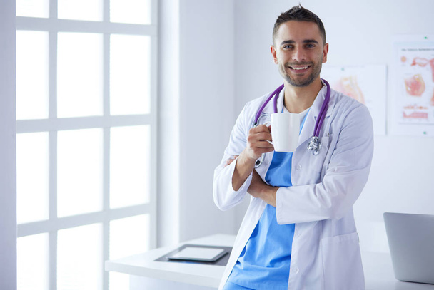 Młody i pewny siebie mężczyzna lekarz portret stojący w gabinecie medycznym. - Zdjęcie, obraz