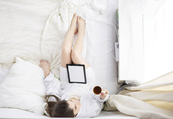Portrait of woman using tablet and drinking coffee while sitting on the bed in the morning - Valokuva, kuva