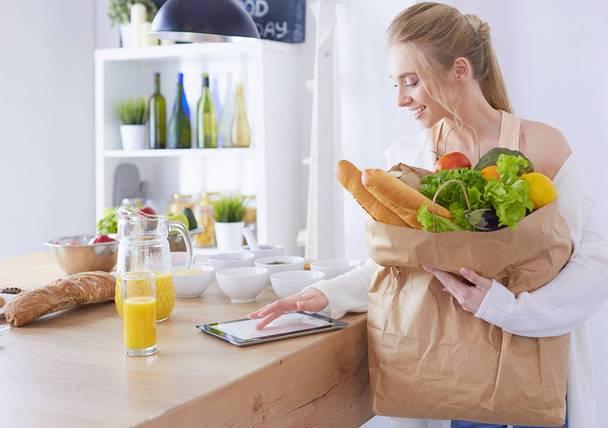 Młoda kobieta trzyma torbę na zakupy z warzywami.Stojąc w kuchni - Zdjęcie, obraz