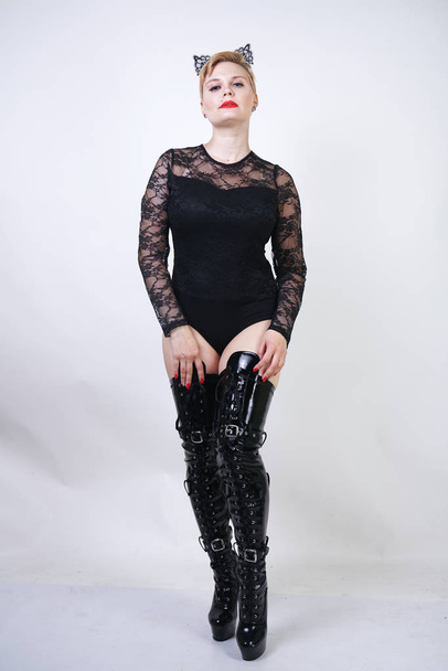 pretty plus size young female with short blonde hair posing on white studio background. hot chubby fashion girl in black lace bodysuit and patent leather thigh high boots standing as cat woman alone. - Foto, immagini