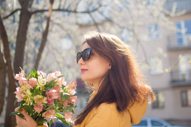Sensual young girl in dark sunglasses has just received a big bouquet of colorful flowers outdoors near the buildings on the background - Photo, Image