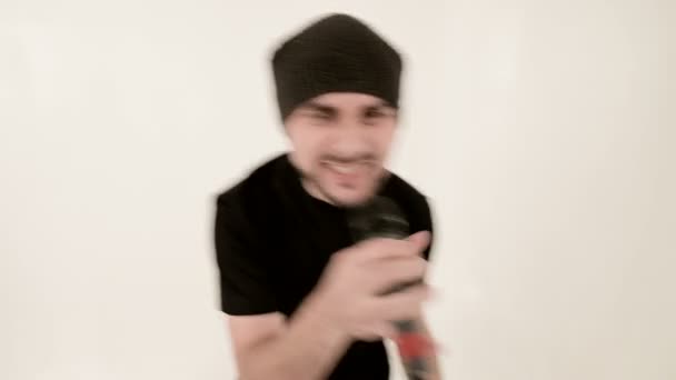 Frontman vocalist rock pop with a stylish beard in black clothes and a hat with a microphone in his hands expressively aggressively singing in the studio against the background of white walls - Πλάνα, βίντεο
