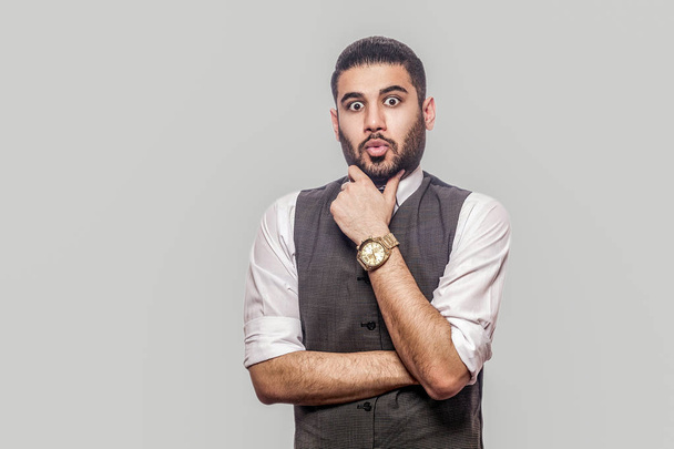 suprised handsome bearded brunette man in white shirt and waistcoat holding his chin and looking at camera with shocked face on gray background. - Foto, Bild