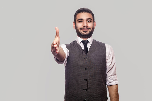 happy handsome bearded brunette man in white shirt and waistcoat looking and smiling at camera while giving hand to handshake and greeting on gray background. - Foto, immagini