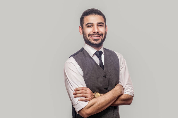 happy glad handsome bearded brunette man in white shirt and waistcoat standing with crossed arms and looking at camera with  toothy smile on gray background  - Foto, Imagem