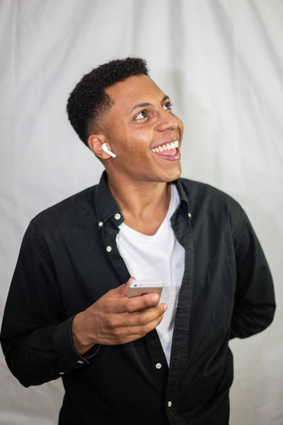 スマートフォンを持って笑う黒いシャツにワイヤレスイヤホンを持つ幸せなアフリカ系アメリカ人の若いハンサムな男 - 写真・画像