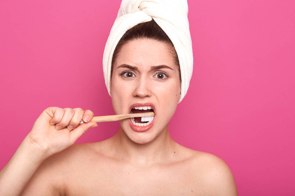 Indoor studio shot of displeased energetic young woman looking directly at camera, brushing teeth intesively with toothbrush, being naked with towel on head, standing isolated over pink background. - Foto, afbeelding
