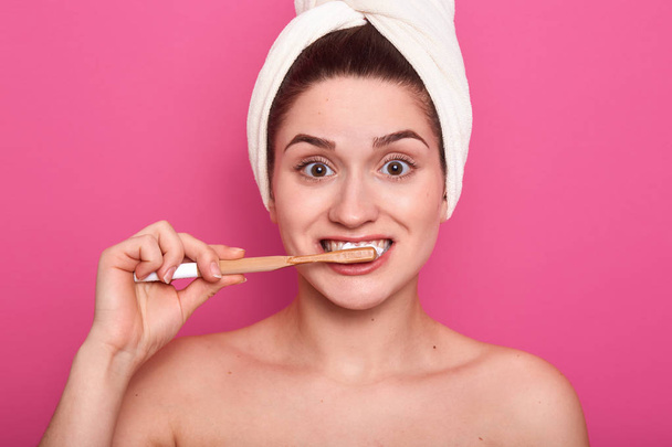 Close up shot of girl with funny facial expression, posing with toothbrush, young female cleans her teeth, standing in bathroom with towel on head, isolated over pink background. Oral hygiene concept. - Photo, image