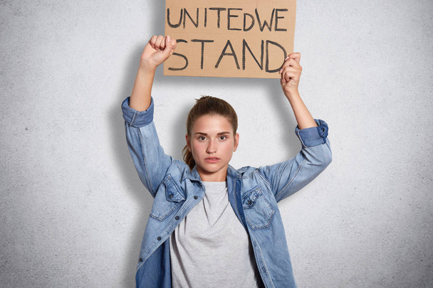 Portrait of militant powerful young feminist showing her fist, holding sign with inscription united we stand in one hand, having decisive facial expression, interested in women social problems. - Photo, image