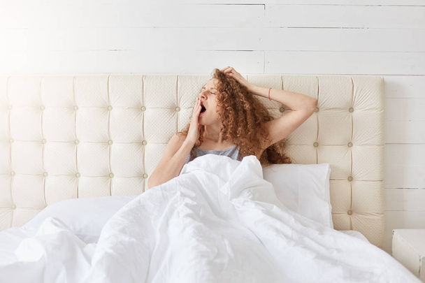 Indoor shot of peaceful attractive young lady relaxing in her bed in morning, yawning, covering her mouth with one hand, having curly hair, enjoying start of new day. People and morning concept. - 写真・画像