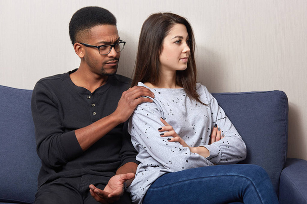 Offended attractive woman sitting back to her husband on sofa in flat, african male touches girl's shoulders and asks to forget him, couple wearing casual outfit. Quarrel and relationship concept. - Foto, imagen