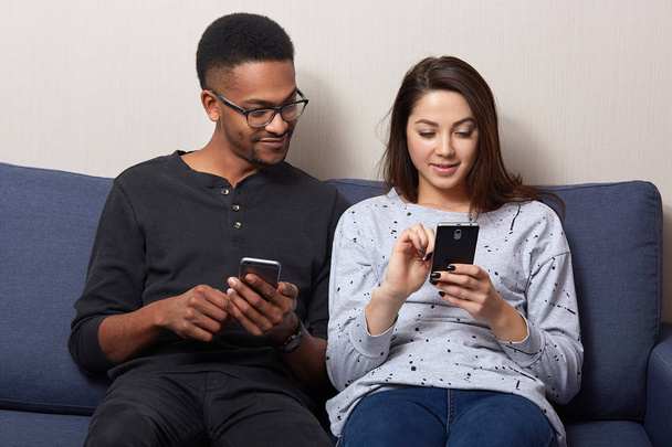 Multicultural couple sitting on sofa at home looking in theirs smart phones, using internet, being happy spending time together, woman showing something in cell phone. People and family time concept. - Foto, Imagen